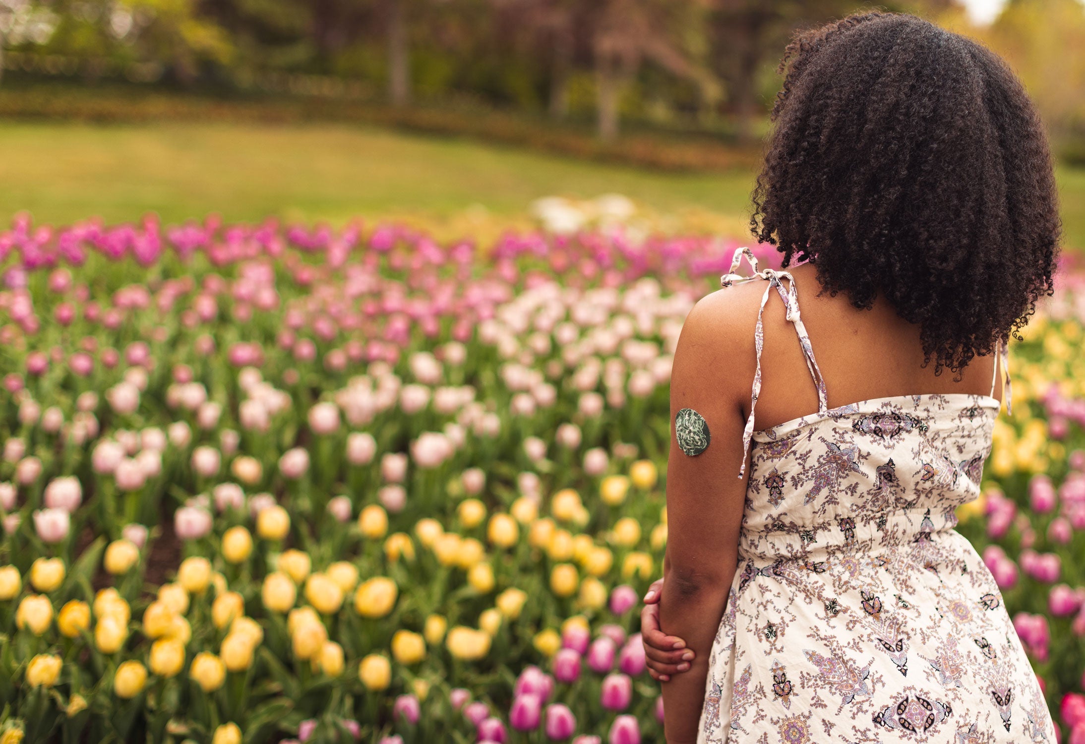 A woman in a floral sundress viewed from behind, standing in a colorful tulip field, with a green floral- patterned round CGM adhesive patch on her upper arm