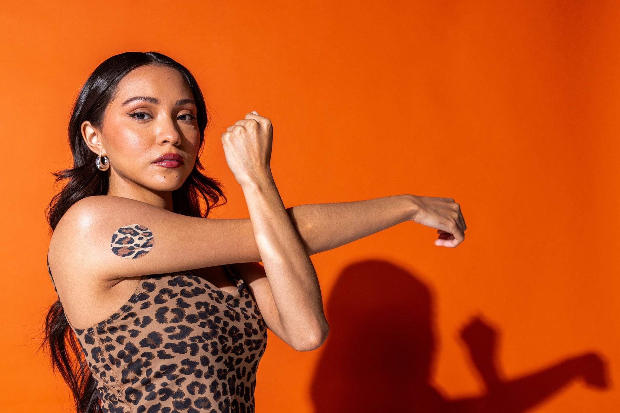 A woman in a leopard-print dress posing dramatically against a bright orange background, with a leopard-print round CGM adhesive patch on her upper arm