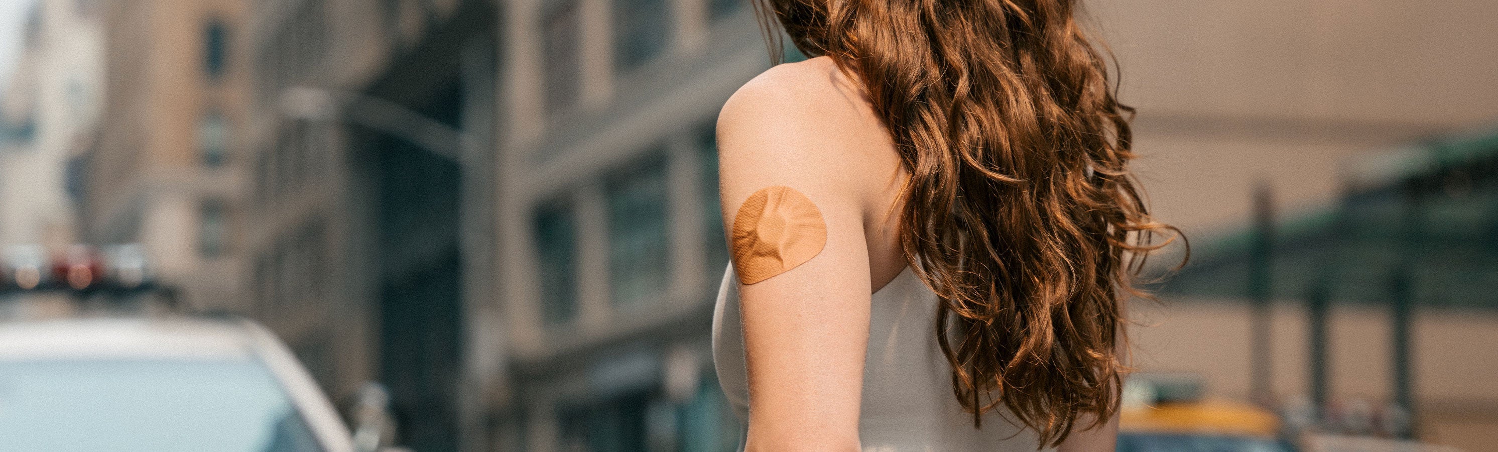 A woman with long auburn wavy hair viewed from behind on a city street, with a tan/nude floral- patterned round CGM adhesive patch on her upper arm
