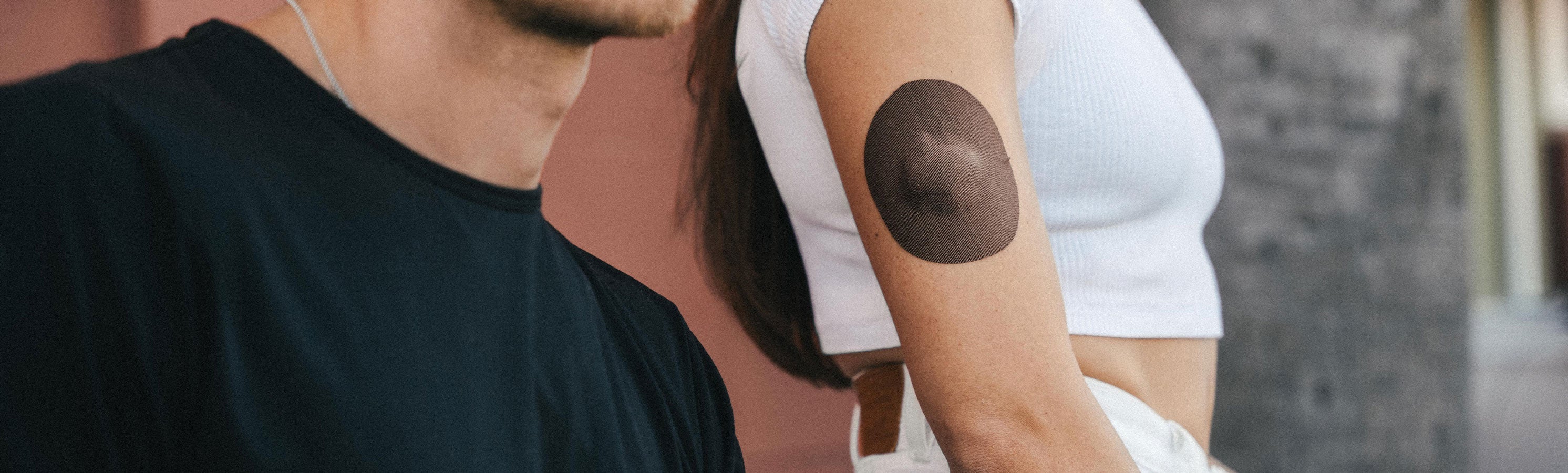 Two people standing close together outdoors; a woman in a white crop top has a dark brown/chocolate round CGM adhesive patch on her upper arm