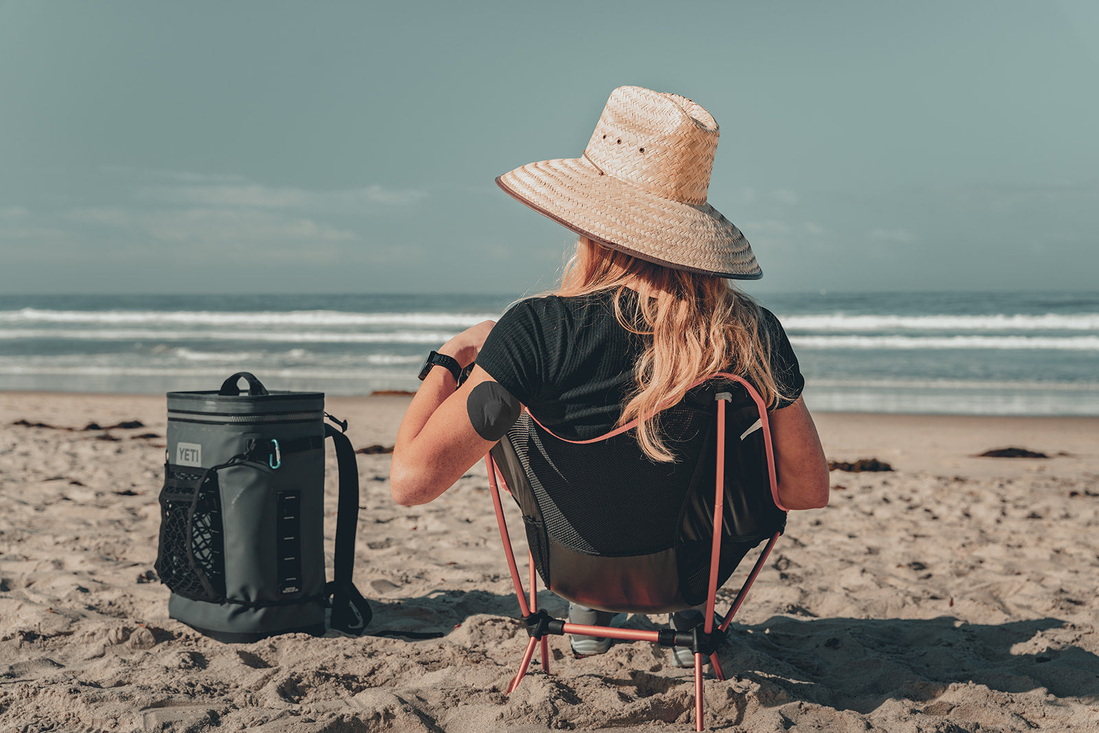 A woman viewed from behind sitting in a folding camp chair on the beach, wearing a wide-brim straw hat and black t-shirt, with a dark round CGM adhesive patch visible on her upper arm; a backpack cooler sits beside her in the sand