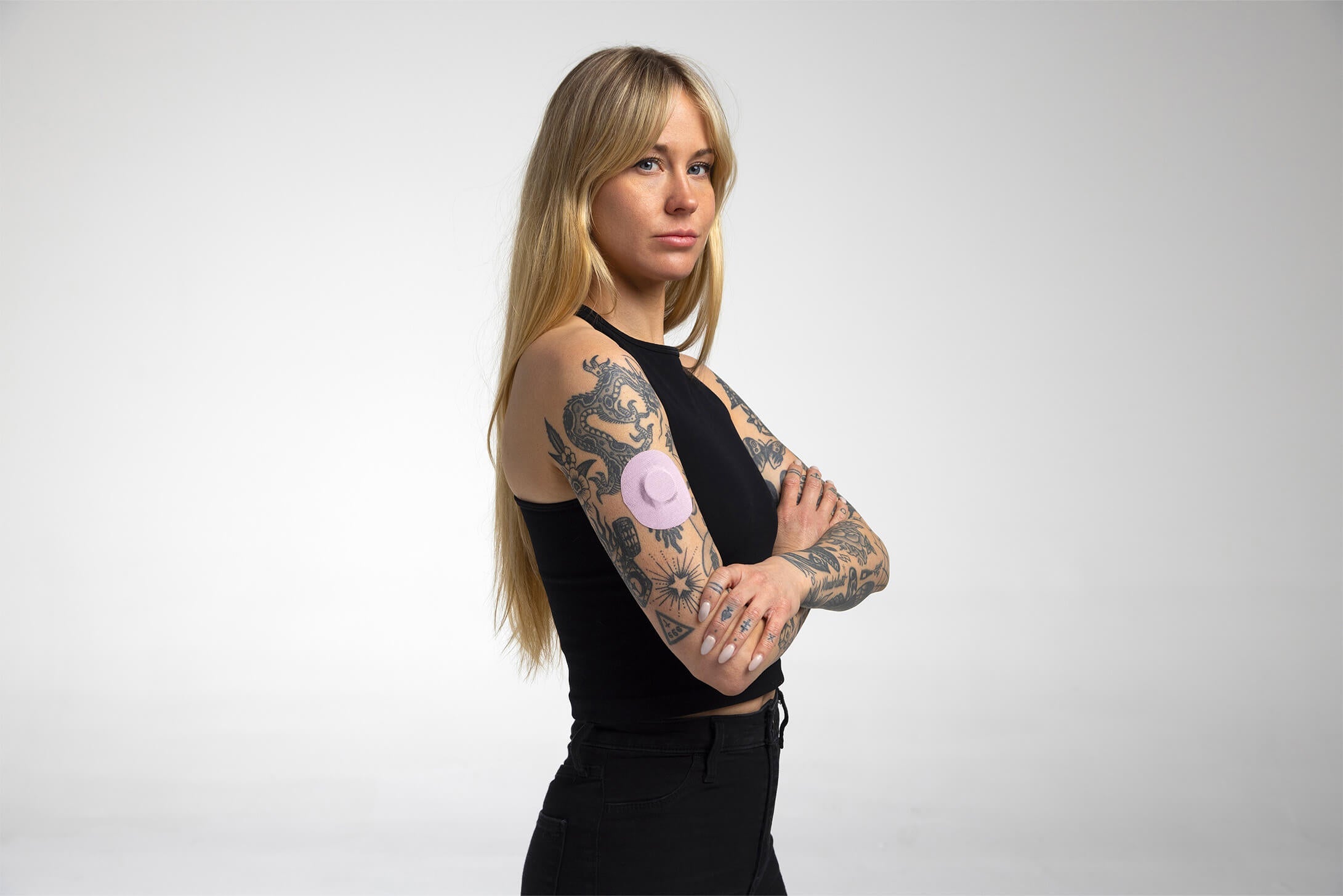 A tattooed blonde woman in a black outfit with arms crossed, looking over her shoulder; a soft lavender round CGM adhesive patch is visible on her forearm