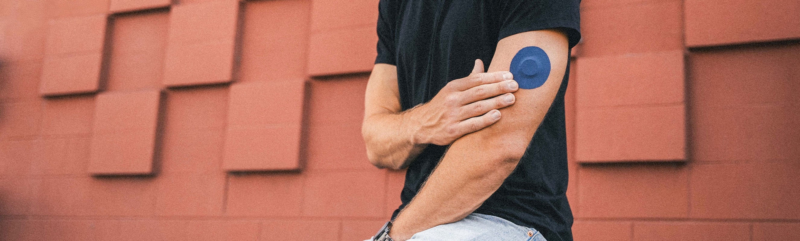 A man in a black t-shirt touching a solid navy blue round CGM adhesive patch on his upper arm, standing against a terracotta brick wall
