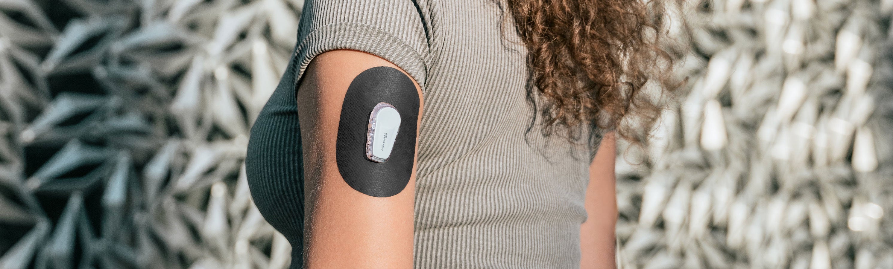 Close-up of a person's upper arm in a sage green ribbed top with curly hair, wearing a solid black rounded-rectangular CGM adhesive patch with a small white sensor visible