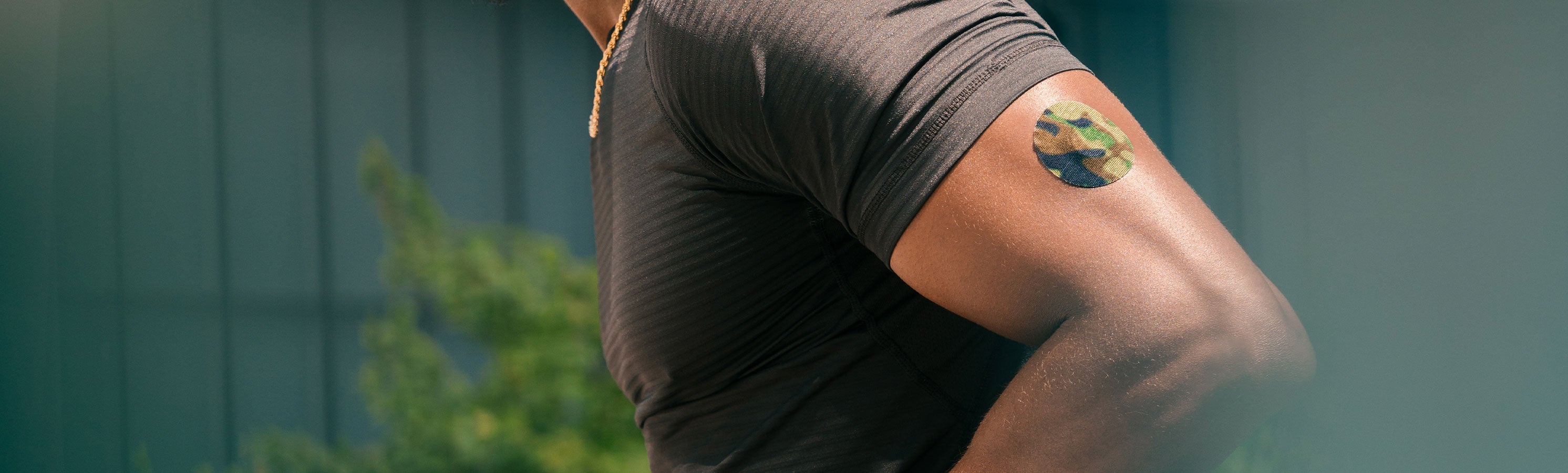 A person in a gray athletic shirt viewed from behind and side, with a camouflage-patterned round CGM adhesive patch on their upper arm
