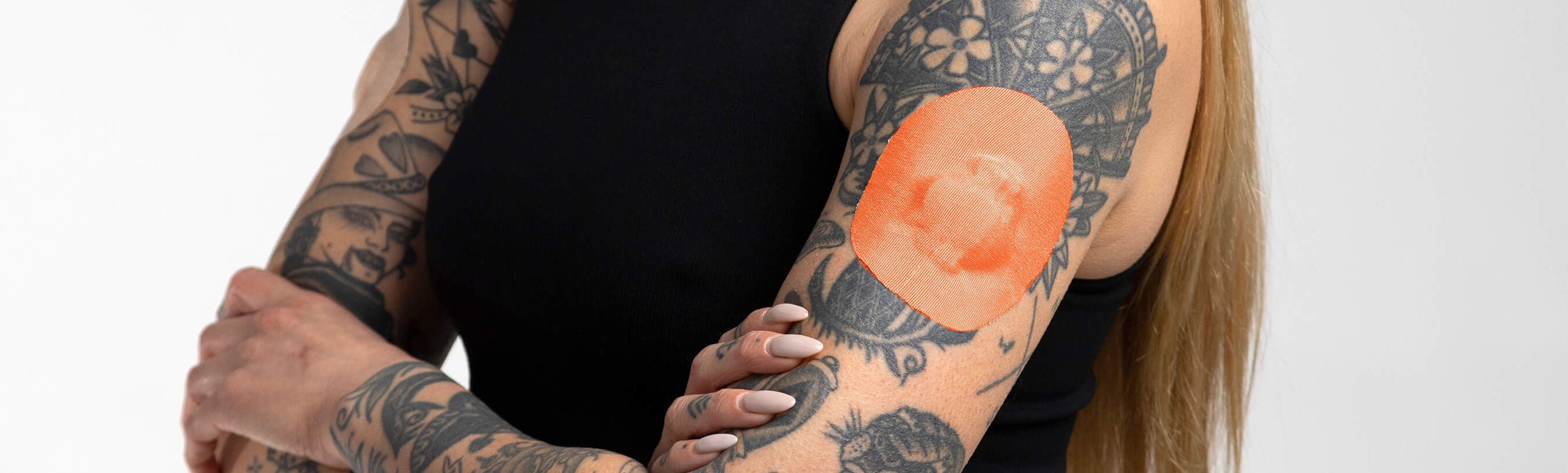 Close-up of two heavily tattooed arms crossed, with an orange-toned round CGM adhesive patch visible on the upper arm
