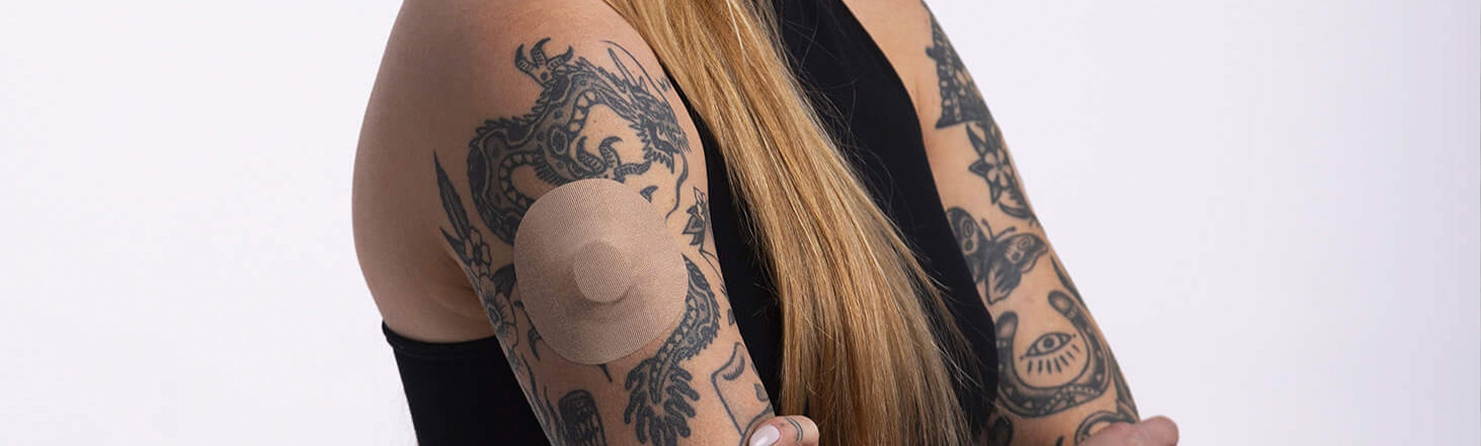 Close-up of a heavily tattooed arm, with a skin-tone beige round CGM adhesive patch on the upper arm; the person has long blonde hair and is wearing a black top