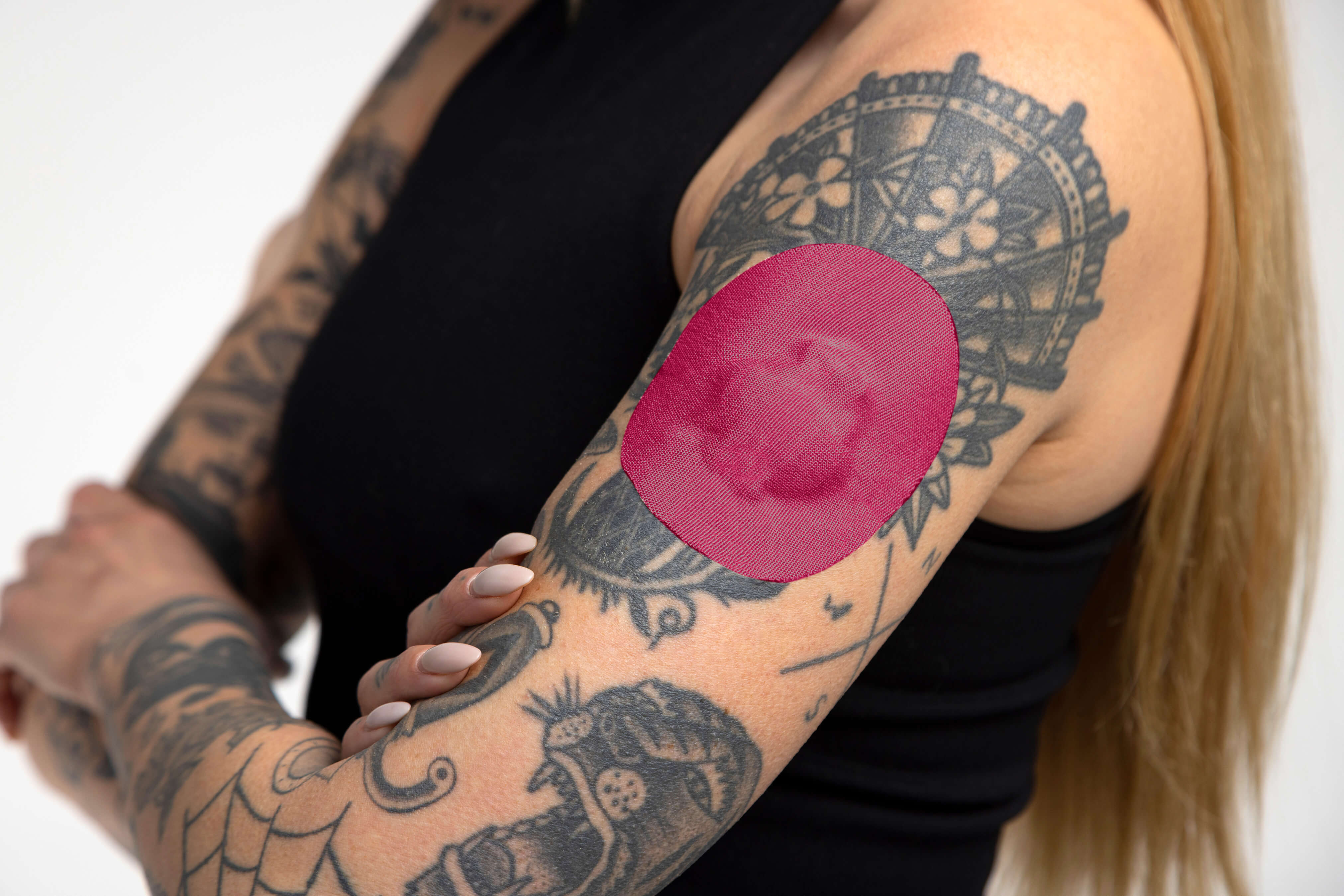 A heavily tattooed woman in a black tank top with arms crossed, showing a large rose-pink round CGM adhesive patch on her upper arm