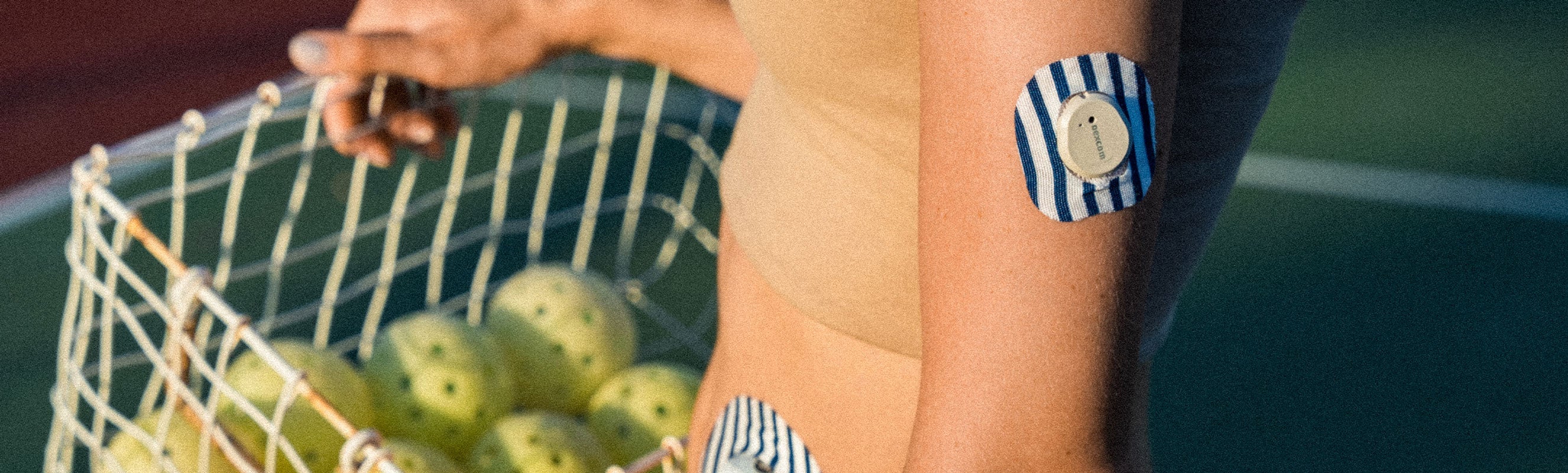 Close-up of a person's torso and arm holding a wire basket filled with yellow-green pickleball balls, with a blue-and-white striped square CGM adhesive patch on their upper arm against a dark teal background
